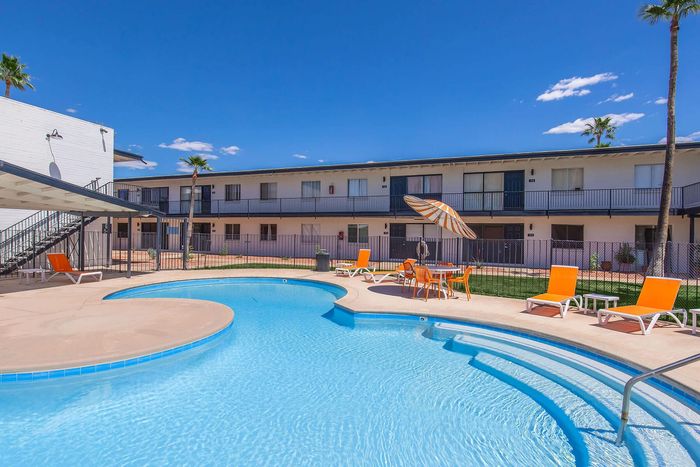 Inviting apartment complex showcasing a sparkling swimming pool with orange lounge chairs and umbrellas set against a bright blue sky.