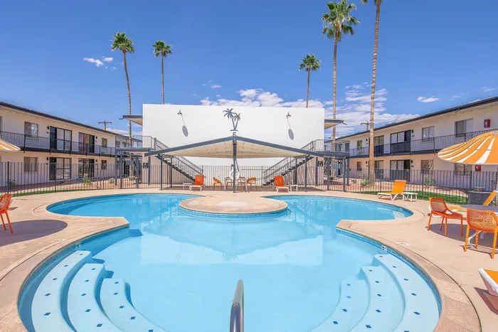 Inviting apartment complex pool area with vibrant orange lounge chairs and umbrellas, surrounded by palm trees and clear blue skies.