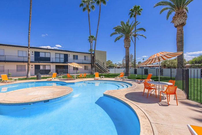 Inviting outdoor pool area with vibrant orange lounge chairs and umbrellas, surrounded by lush palm trees and a well-maintained apartment building under a clear blue sky.