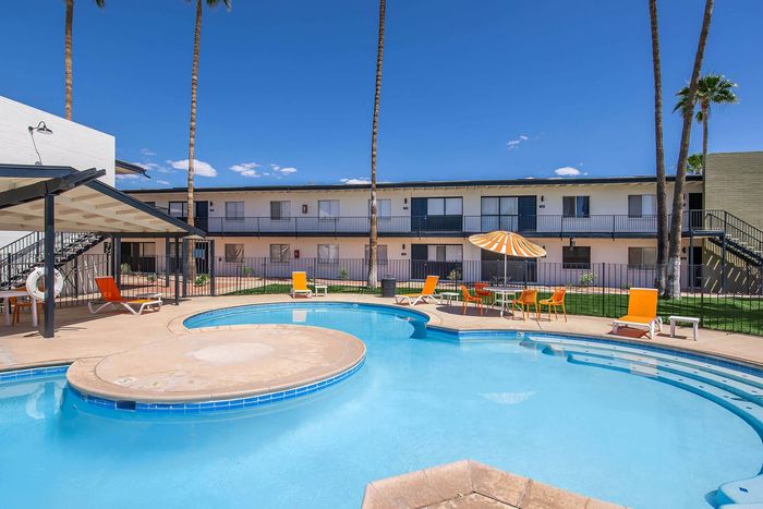 Vibrant courtyard view featuring a sparkling swimming pool with stylish orange loungers and umbrellas, surrounded by modern apartment buildings and lush palm trees.