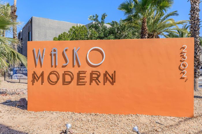 Wasko Modern sign with bold lettering against a vibrant orange background, surrounded by desert landscaping and palm trees, offering a modern living space.