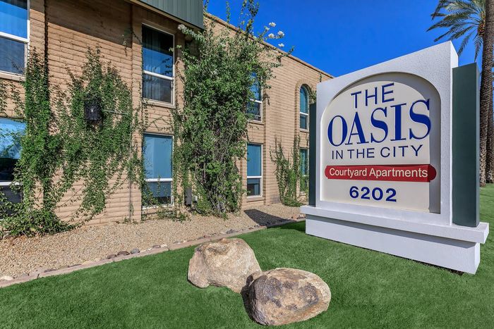 Charming courtyard apartment complex with lush greenery and a prominent sign reading 'The Oasis in the City' against a clear blue sky.