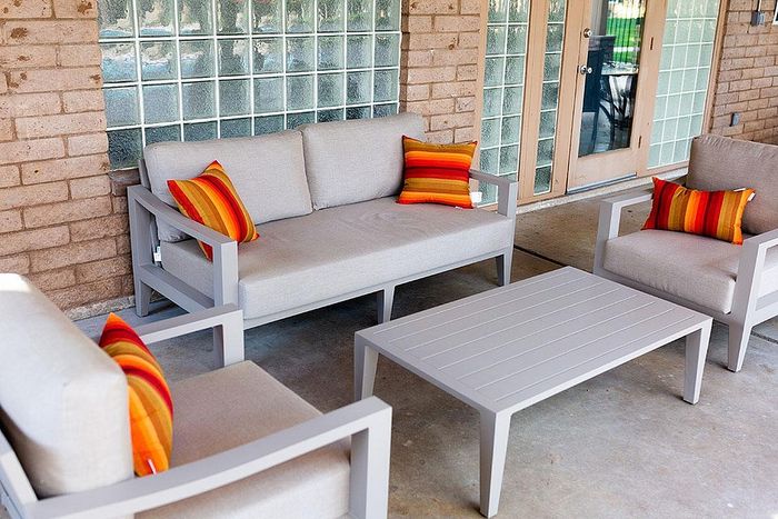 Chic outdoor patio with modern gray couches, vibrant orange striped pillows, and a sleek coffee table against a textured brick backdrop.
