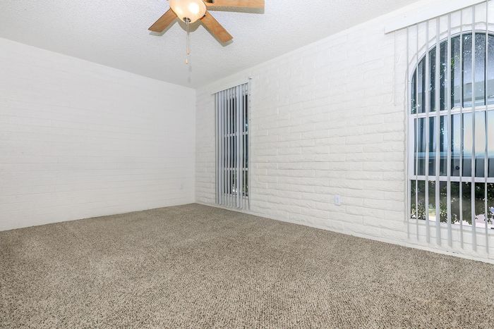 Bright and spacious bedroom with plush carpet, elegant brick walls, and a ceiling fan, featuring ample natural light through large windows with blinds.