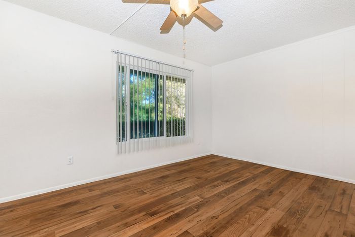 Bright room with elegant hardwood flooring and large window, featuring ceiling fan and vertical blinds, offering a serene view of lush greenery.