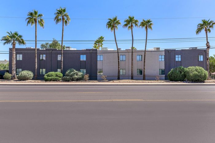 Modern apartment building with sleek gray facade, lined with tall palm trees and lush greenery, under a bright blue sky.