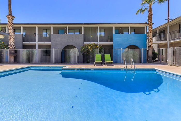 Modern apartment complex with a sparkling swimming pool and relaxing lounge chairs, surrounded by palm trees and contemporary architecture.