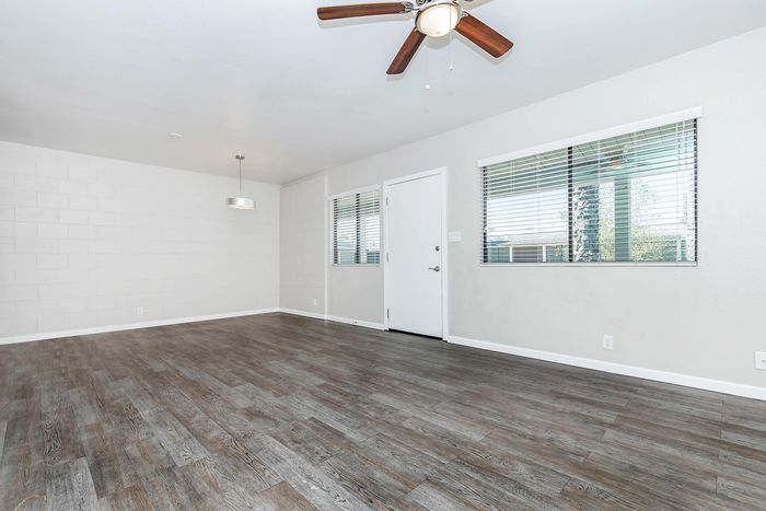 Bright and airy living space with elegant wood flooring, modern ceiling fan, and ample natural light from large windows.