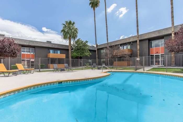 Inviting apartment complex with a sparkling pool, surrounded by elegant lounge chairs and tall palm trees against a clear blue sky.