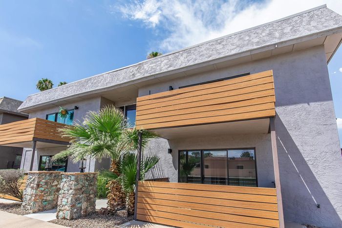 Modern apartment exterior with sleek wood paneling, large glass windows, and lush palm landscaping against a vibrant blue sky.