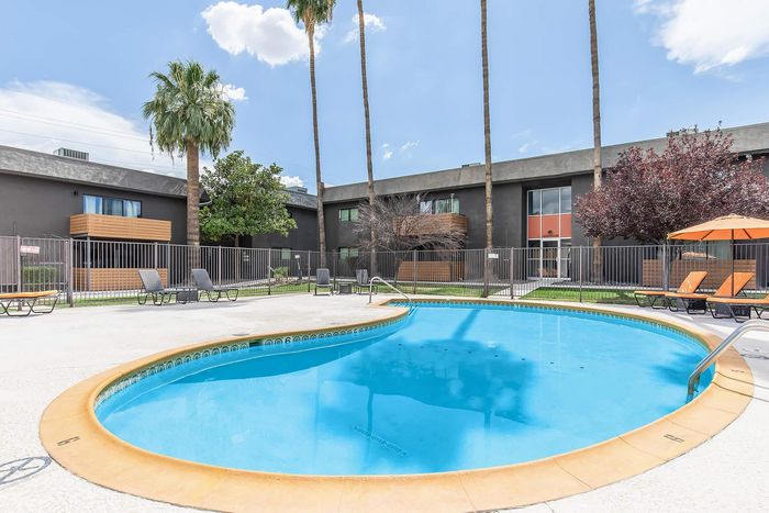 Inviting apartment complex featuring a bright outdoor pool, sun loungers, and lush tropical palm trees against a clear blue sky.