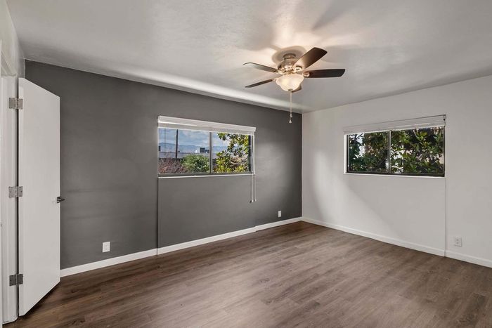 Bright and airy bedroom with sleek laminate flooring, two large windows, and modern ceiling fan, perfect for relaxation.