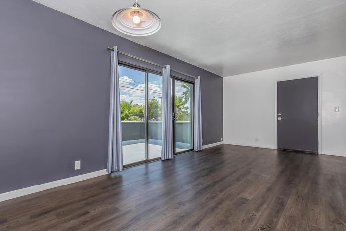 Modern living room with sleek hardwood floors, large sliding glass doors, and a soft gray accent wall, opening to a bright balcony view.
