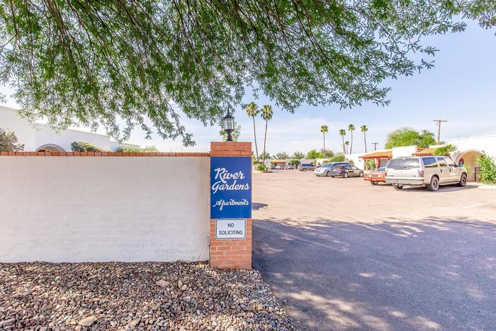Charming entrance to River Gardens Apartments, featuring a welcoming sign and a well-maintained parking area under lush trees.