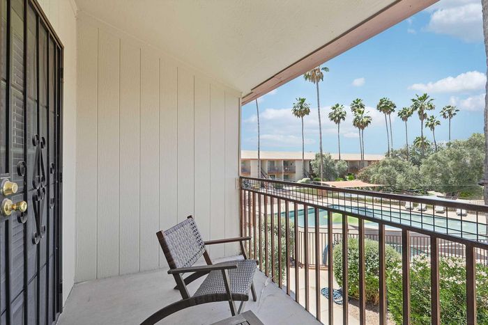 Charming balcony view featuring a cozy seating area overlooking a lush courtyard with a sparkling pool and tall palm trees under a clear blue sky.