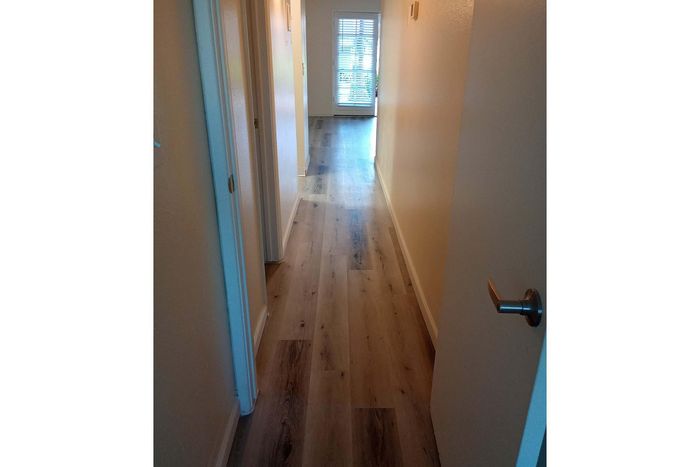 Inviting hallway with elegant wood flooring leading to a sunlit room with large windows and blinds.