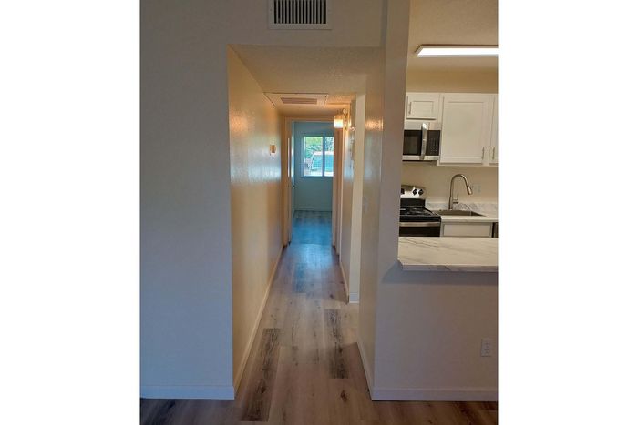 Inviting apartment hallway with modern wood flooring, leading to a sunlit window and adjacent to a contemporary kitchen featuring sleek white cabinetry and stainless steel appliances.