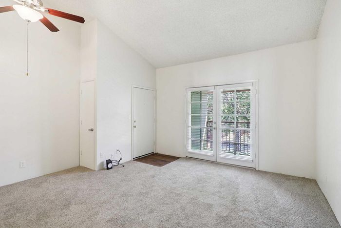 Bright and spacious living room featuring plush carpeting, ceiling fan, and expansive windows leading to a serene balcony view.