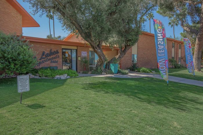 Inviting entrance of a brick apartment complex surrounded by lush greenery and colorful 'Now Leasing' banners, offering a welcoming atmosphere.