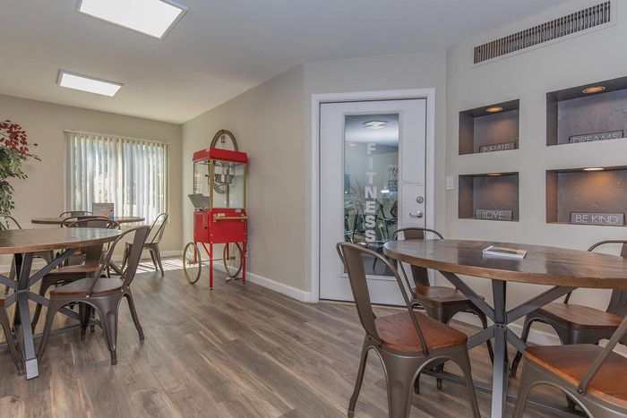 Inviting communal space with round wooden tables, metal chairs, and a vibrant red popcorn machine adds a playful touch. A door labeled 'Fitness' leads to a gym area, surrounded by stylish wall niches with inspirational words.