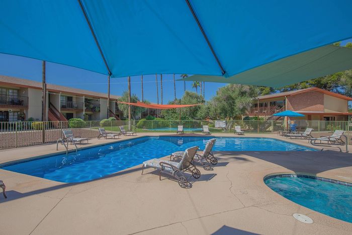 Inviting apartment community pool area with lounge chairs, vibrant blue canopy, and lush greenery under a clear sky.