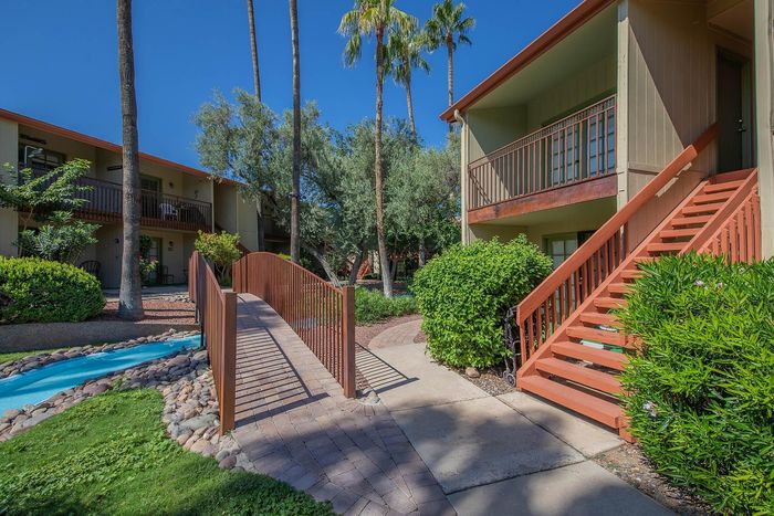 Charming apartment exterior with lush greenery, a wooden bridge over a serene creek, and a sunny balcony view.