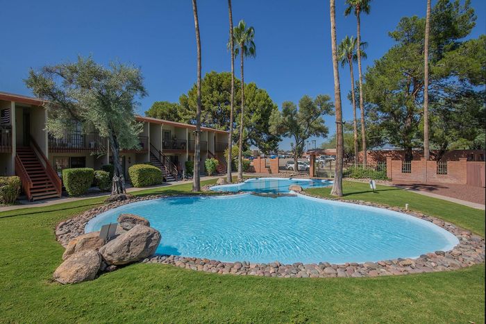 Charming apartment complex featuring lush landscaping and a sparkling outdoor pool surrounded by tall palm trees.
