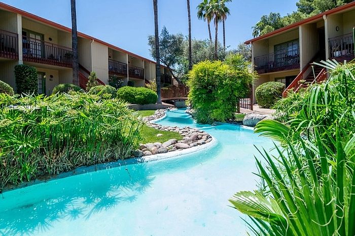 Tranquil apartment complex surrounded by lush greenery and a winding turquoise stream, providing a serene oasis.