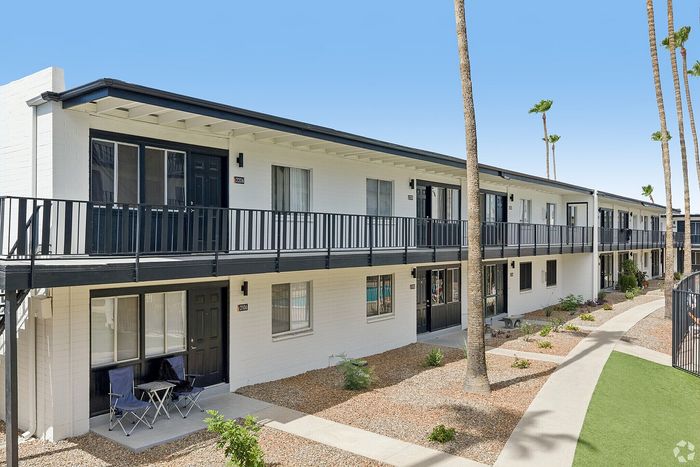 Charming two-story apartment complex with modern balcony railings, surrounded by well-maintained landscaping and tall palm trees under a bright blue sky.