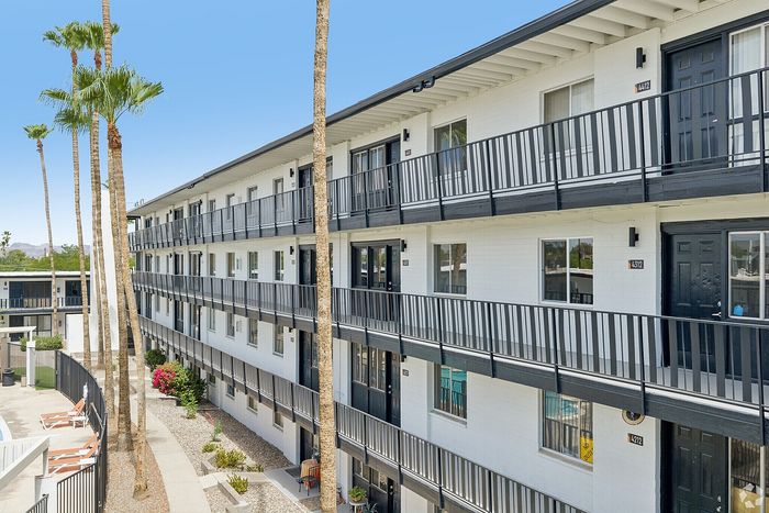 Modern three-story apartment building with sleek black balconies, lined with tall palms and vibrant landscaping, offering a resort-style ambiance.