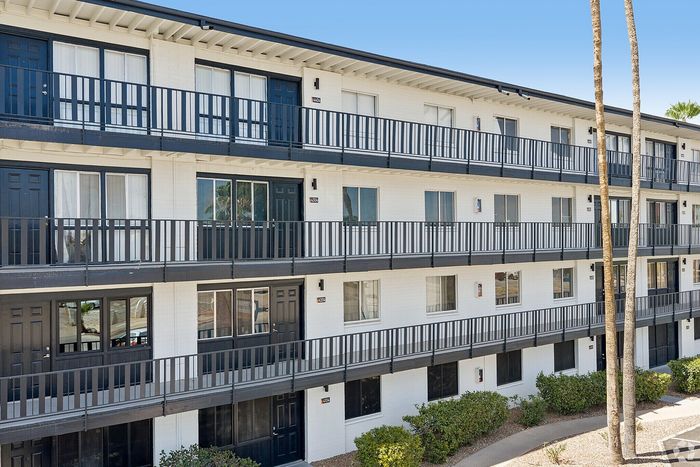 Contemporary apartment building exterior with sleek black railings, large windows, and lush greenery, under a clear blue sky.