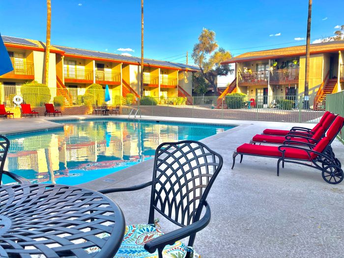 Charming apartment complex with inviting pool, vibrant red lounge chairs, and sunlit balconies surrounded by lush greenery.