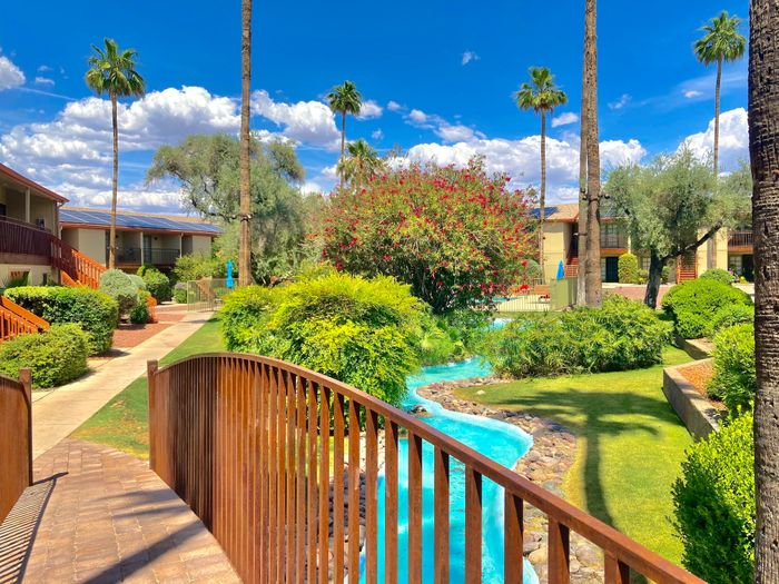 Charming apartment complex with lush landscaping, vibrant gardens, and a picturesque footbridge over a small stream under a bright blue sky.