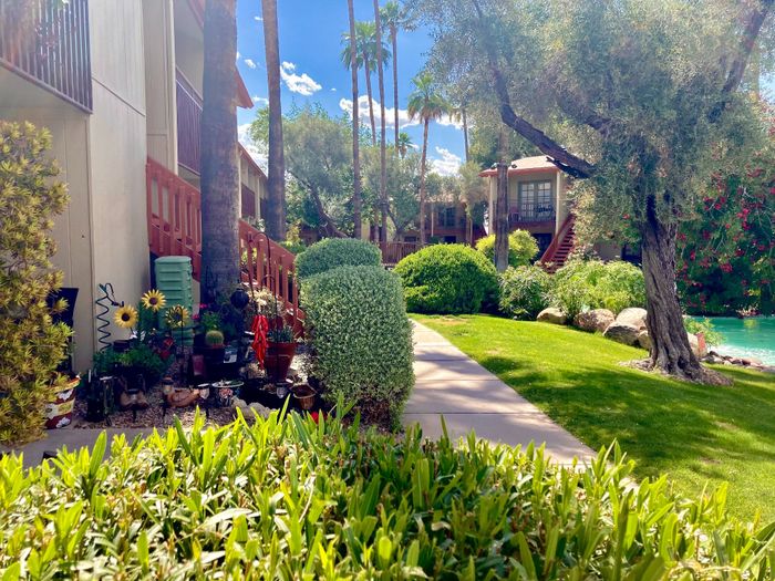 Lush and serene apartment complex with vibrant gardens, mature trees, and a peaceful pathway under a sunny blue sky.