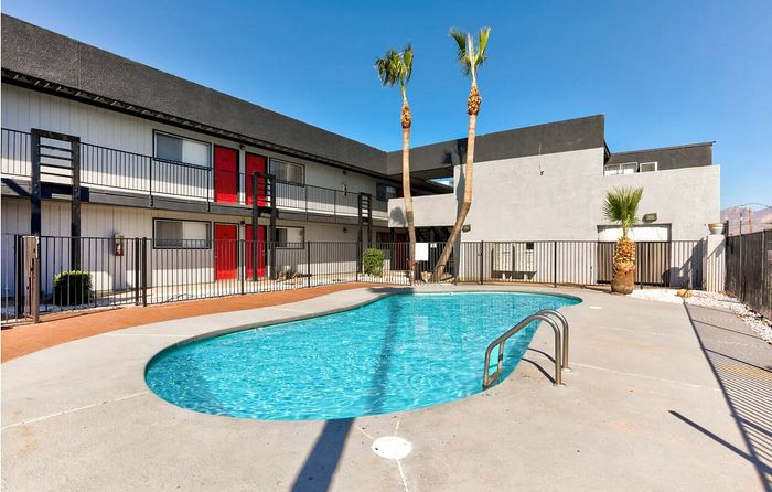 Modern apartment complex with vibrant red doors, featuring a refreshing outdoor swimming pool and tropical palm trees under a clear blue sky.