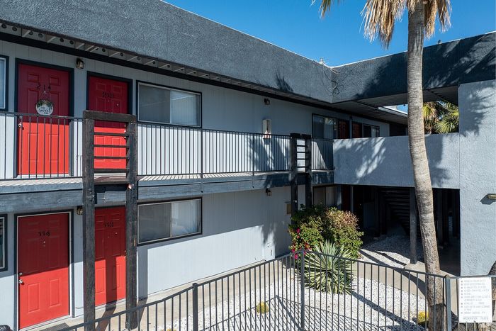 Charming apartment complex featuring vibrant red doors, lush greenery, and a welcoming second-floor walkway under clear blue skies.