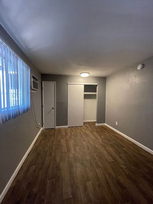 Spacious room with elegant wood flooring, soft gray walls, a modern light fixture, and large window blinds for ample natural light.
