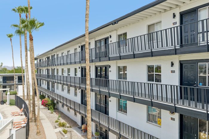 Charming multi-story apartment building with sleek black railings and vibrant landscaping, featuring sunlit balconies and inviting poolside chairs.