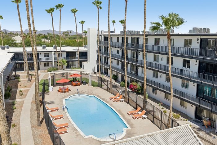 Modern apartment complex featuring a sparkling swimming pool surrounded by palm trees and stylish lounge chairs, with a breathtaking mountain view in the background.