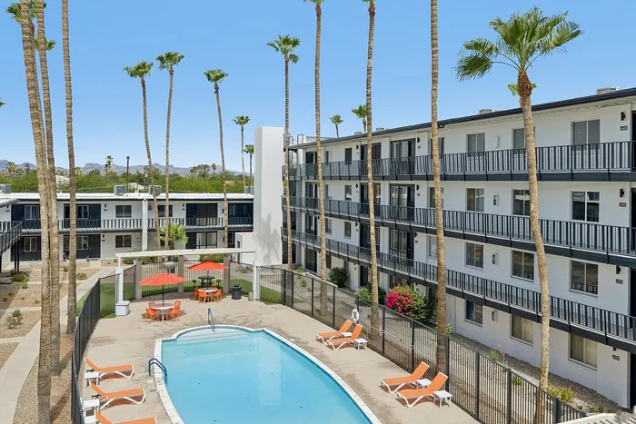 Modern apartment complex with balconies, overlooking a refreshing pool area surrounded by palm trees and vibrant landscaping.