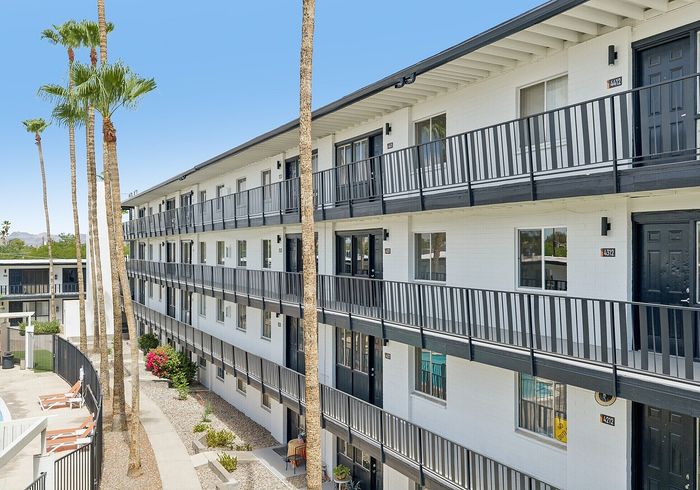 Modern apartment complex with sleek black railings and sunlit balconies, featuring a tranquil outdoor setting with palm trees and a swimming pool area.