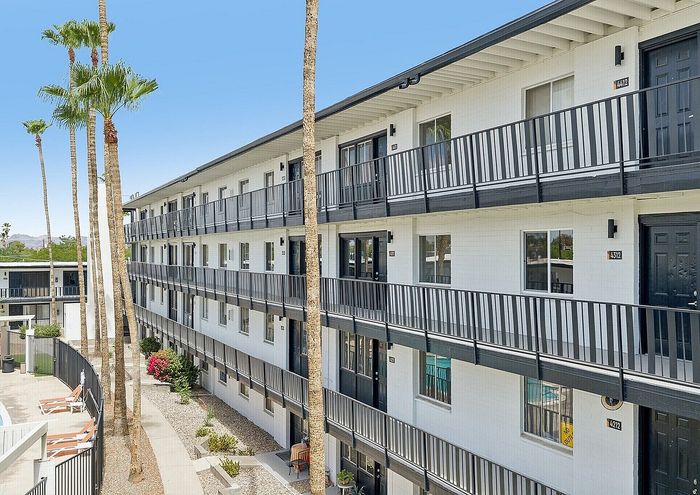 Modern apartment building with multiple floors, sleek black railings, and a vibrant courtyard featuring lush palm trees and a sparkling pool.