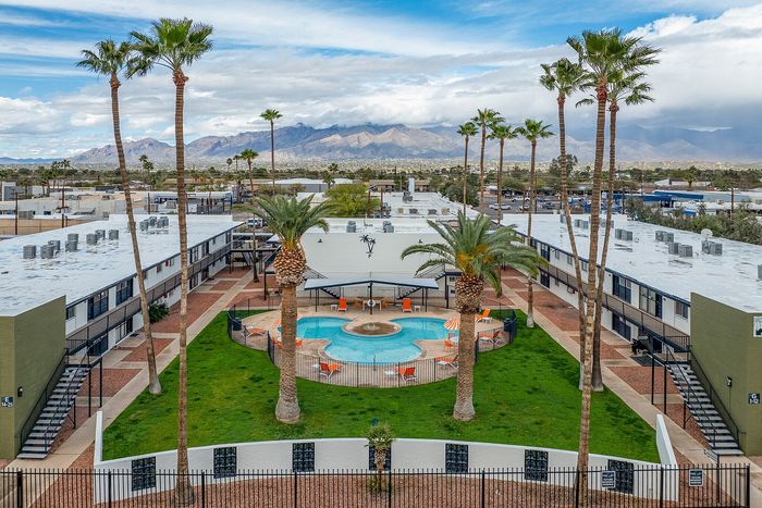 Charming apartment complex featuring a central circular pool surrounded by lush greenery and tall palm trees, set against a backdrop of picturesque mountains and a vibrant blue sky.