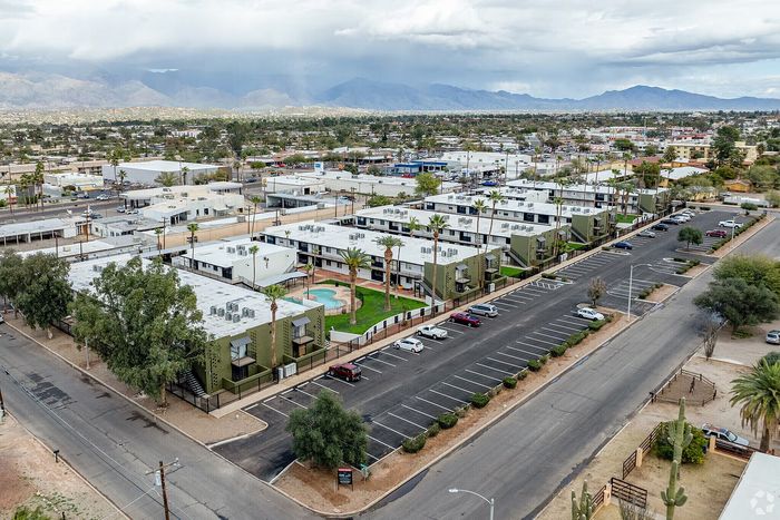 Expansive aerial view of modern apartment complex featuring lush green spaces, palm trees, and ample parking, set against a backdrop of scenic mountains.