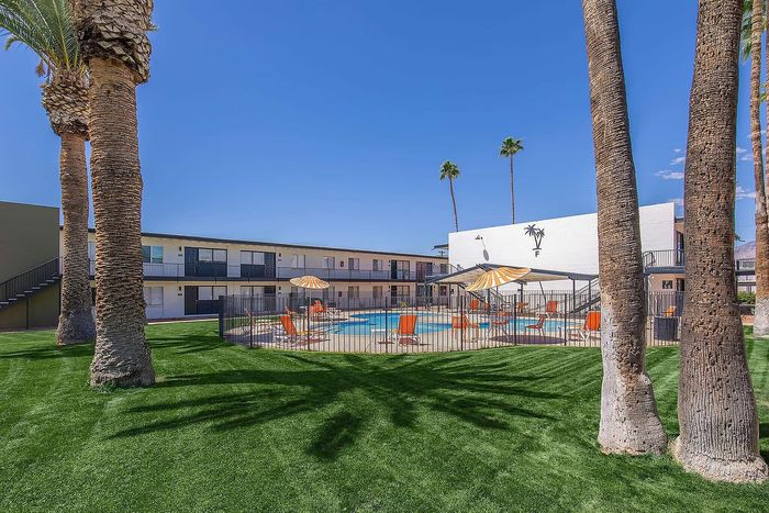 Vibrant apartment complex featuring a sparkling pool, sun loungers, and palm trees against a clear blue sky.