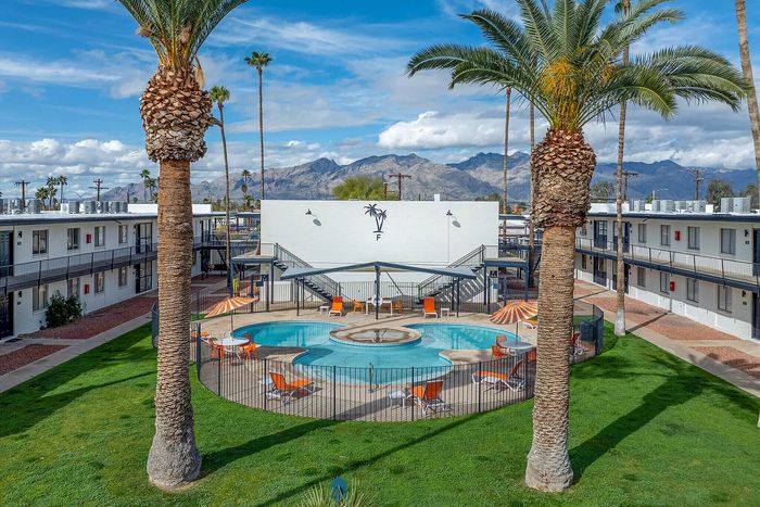 Charming apartment complex with a vibrant courtyard featuring a circular pool surrounded by palm trees and orange sun loungers, set against a stunning mountain backdrop.