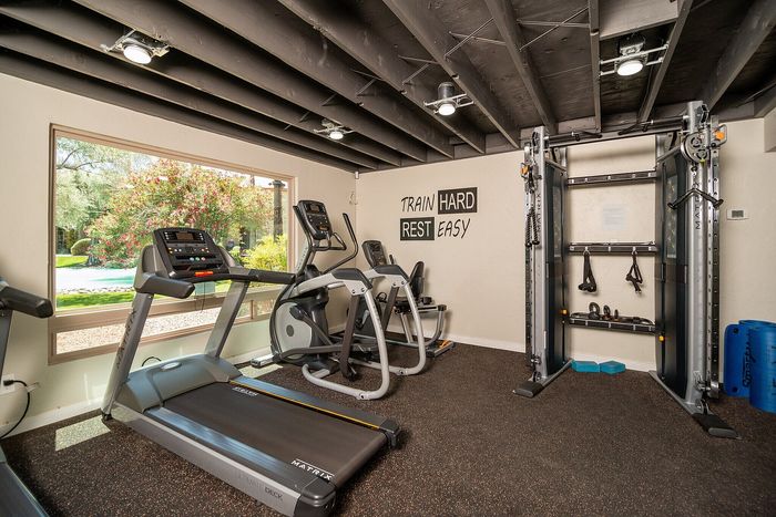 Modern fitness room with state-of-the-art gym equipment, large window providing natural light and a motivational phrase on the wall: 'Train Hard Rest Easy.'