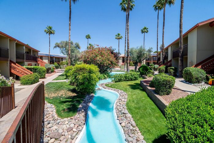 Lush apartment courtyard with tranquil blue stream, surrounded by manicured landscaping and palm trees, creating a serene living environment.