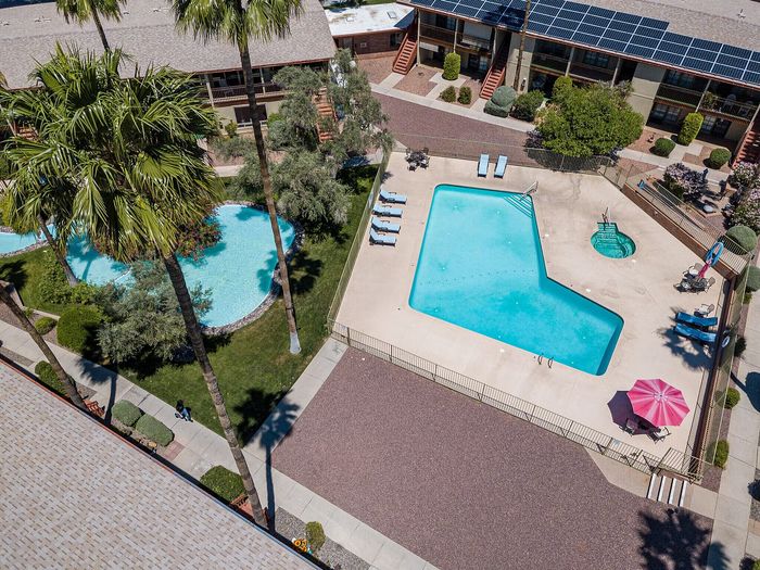 Aerial view of a vibrant apartment complex with a large, uniquely shaped swimming pool, surrounded by lush palms and greenery, offering an inviting oasis atmosphere.