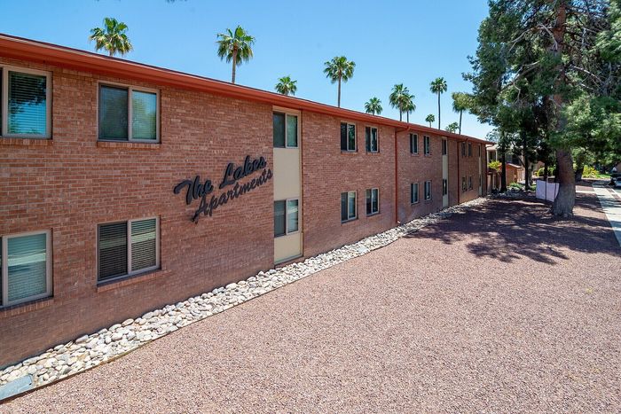 Charming brick exterior of The Lakes Apartments surrounded by palm trees and a serene, landscaped setting.